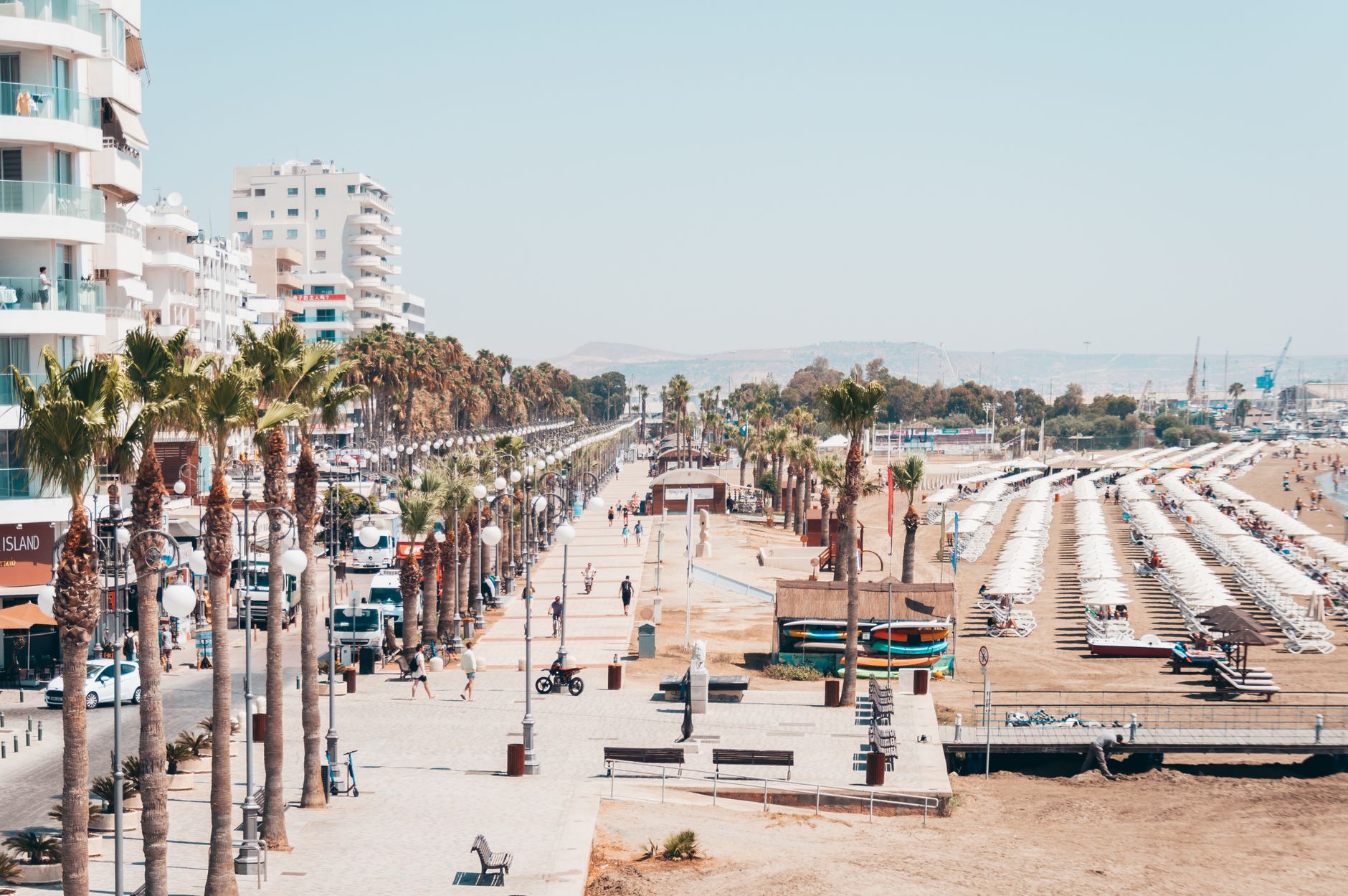 A bustling coastal promenade lined with palm trees, adjacent to a sunny beach with umbrellas and lounge chairs.