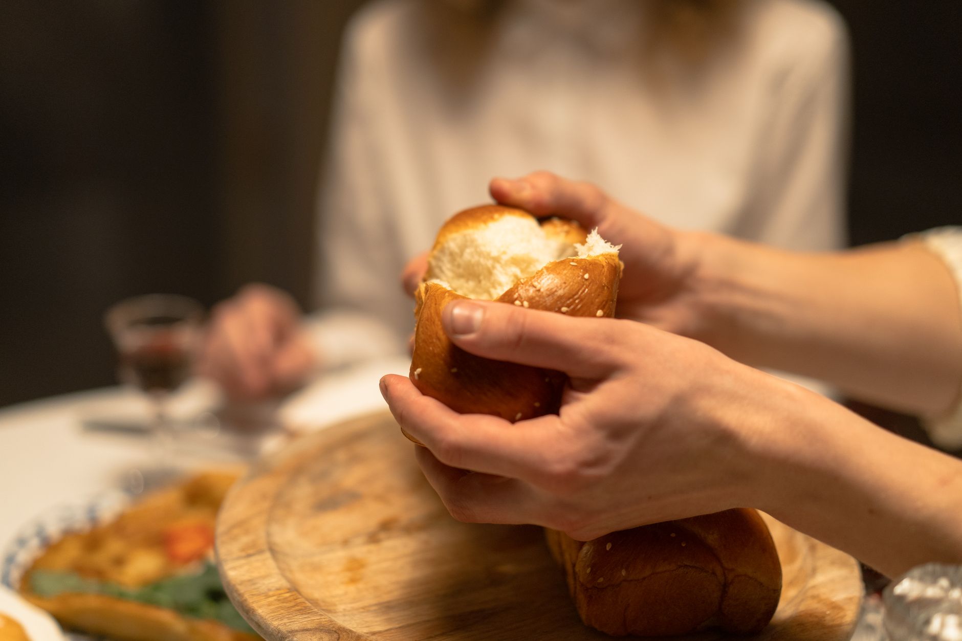 Close-up of hands breaking challah bread at a festive indoor dinner setting.